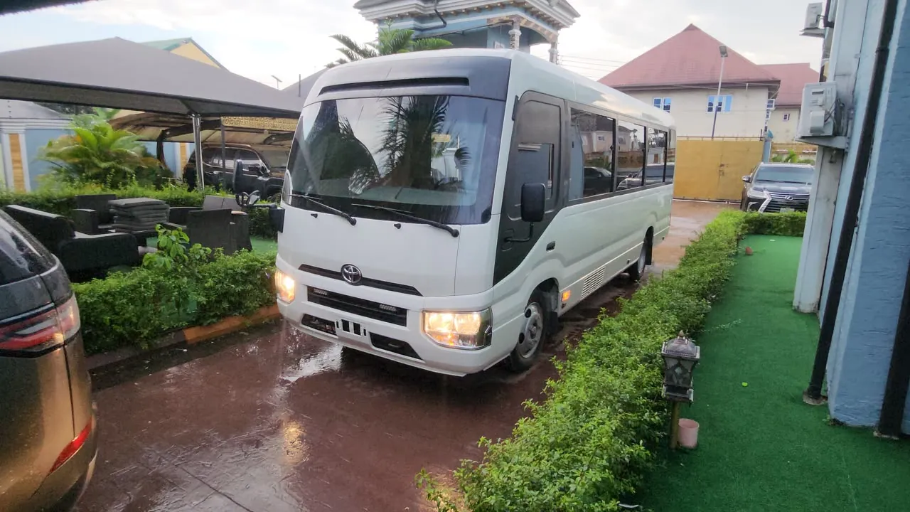 White Toyota Coaster bus with headlights on in a Lagos compound, front angle